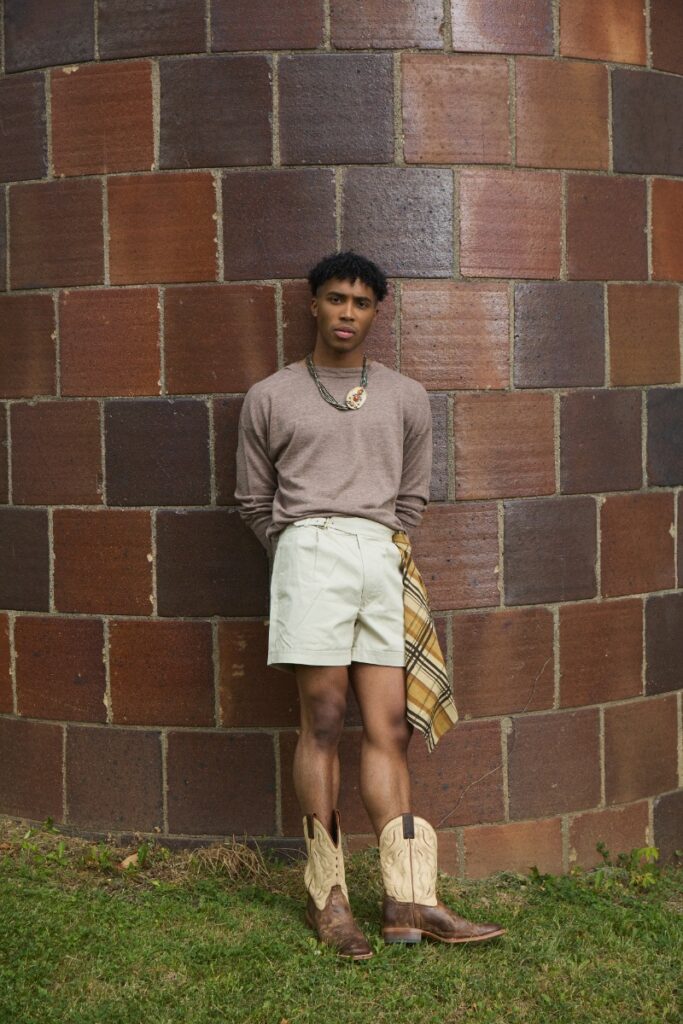 A man leaning against a brick silo wall in a long-sleeve knit shirt, cream shorts, a plaid hip scarf, and two-tone cowboy boots.