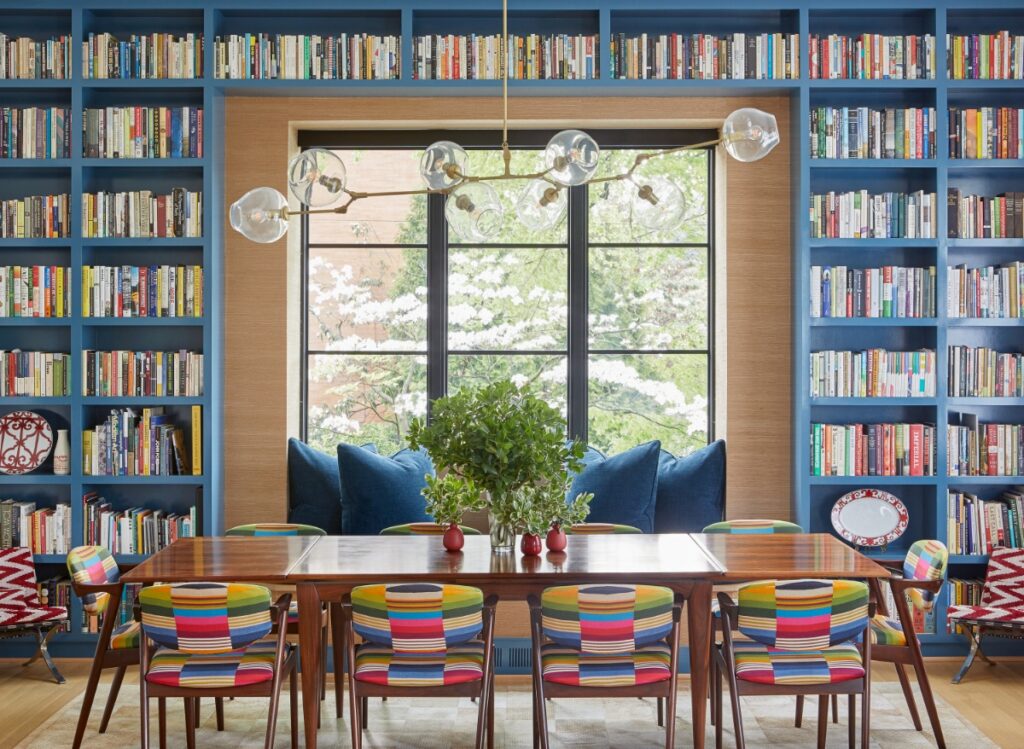 A dining room with built-in blue floor-to-ceiling bookshelves filled with books surrounding a large window, a dark wood table with colorful striped mid-century style chairs, and a modern brass cluster chandelier.