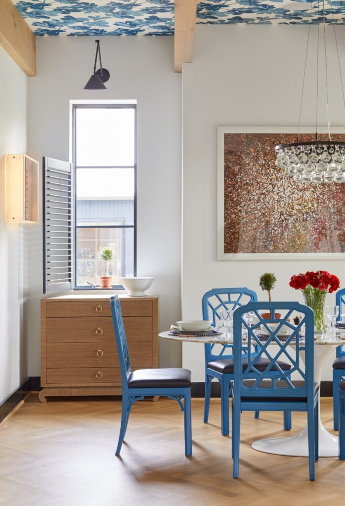 A bright breakfast nook with exposed ceiling beams, blue lattice-back dining chairs around a white pedestal table, a wooden dresser near a window with shutters, and a ceiling accented with blue floral wallpaper.