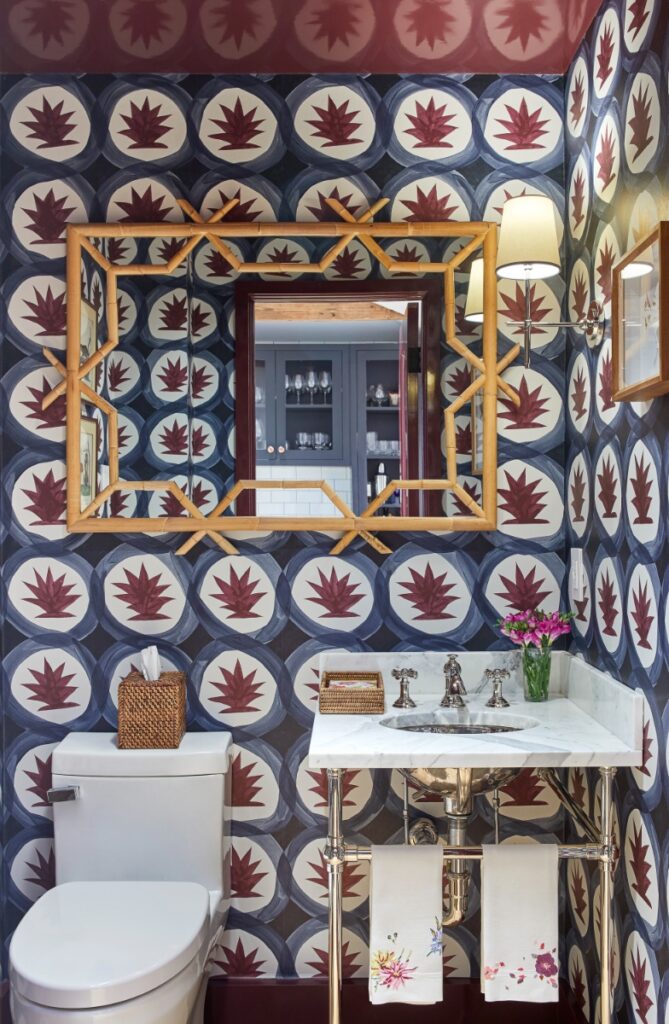 A powder room styled with bold graphic blue and maroon botanical wallpaper, a dark reddish-purple painted ceiling, a bamboo-framed wall mirror, and a marble-topped metal console sink.