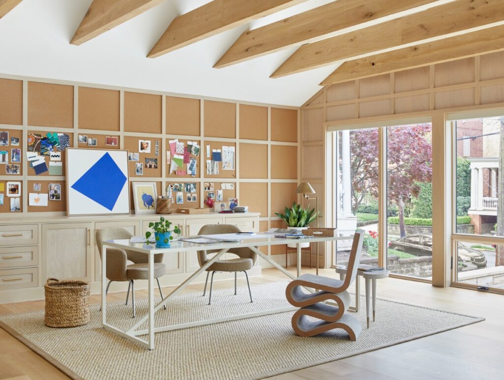 A spacious home office with vaulted ceilings, exposed wood beams, a grid-paneled corkboard wall displaying swatches and photos, a large white desk with beige armchairs, and floor-to-ceiling windows looking outdoors.