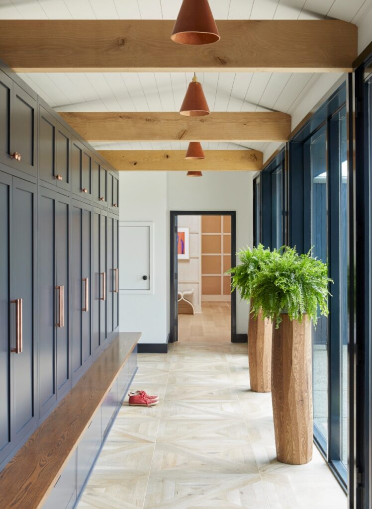 A sunlit hallway or mudroom featuring a row of dark blue cabinetry with copper hardware, built-in bench seating, wood ceiling beams, pendant lamps, and large glass windows overlooking greenery.