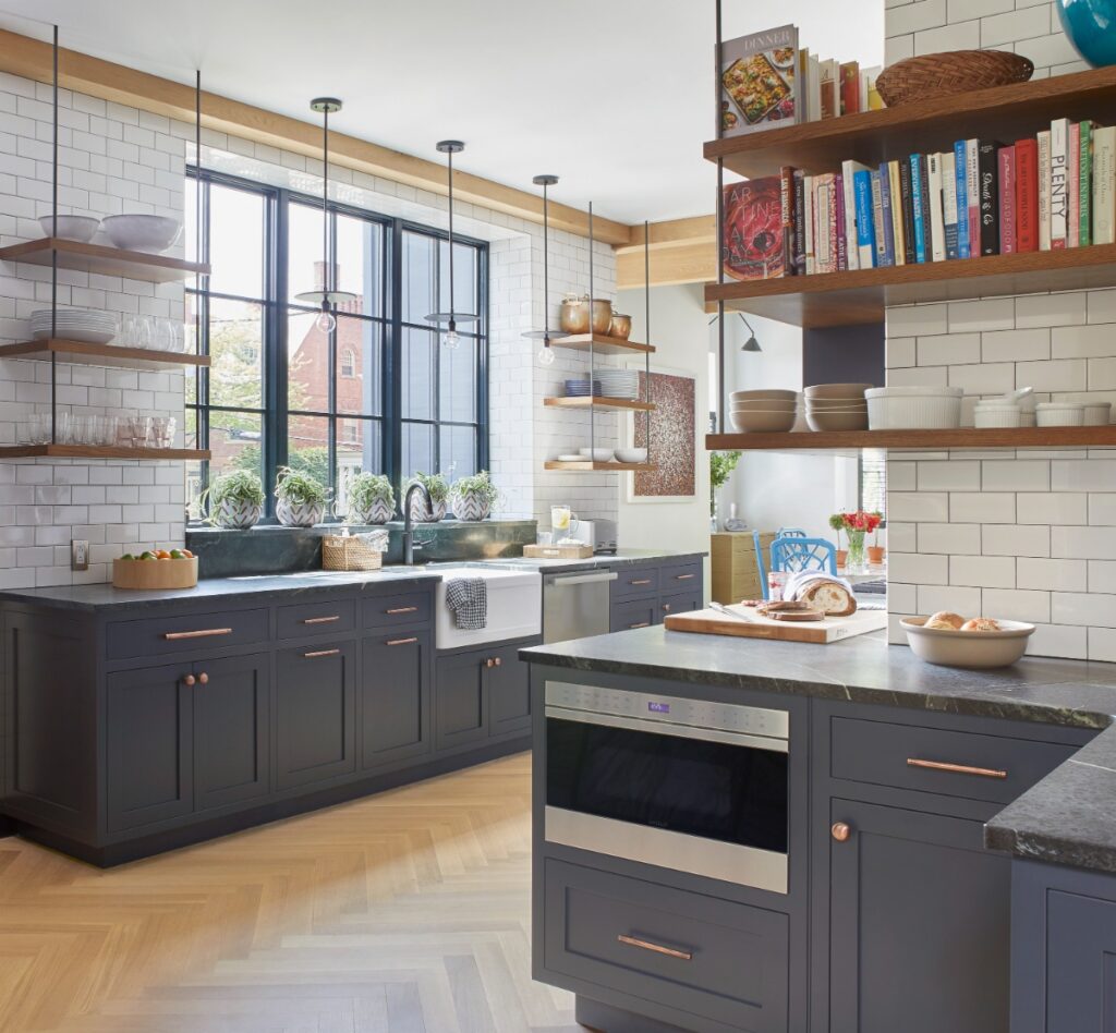 A modern kitchen with dark blue-gray cabinets, copper hardware, dark countertops, white subway tile backsplash, open floating wood shelves displaying cookbooks and dishware, and a large window above the sink.