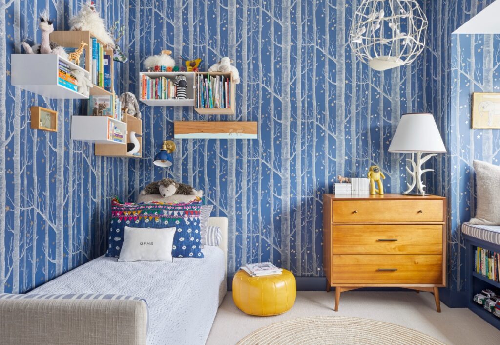 A child's bedroom featuring blue and white tree-patterned wallpaper, a twin bed with colorful patterned throw pillows, a yellow leather pouf ottoman, mid-century wood furniture, and floating wall shelves filled with books and toys.