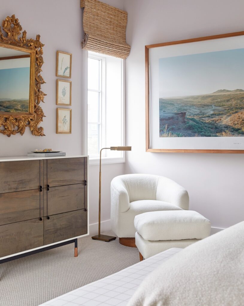 A cozy bedroom seating corner with light lavender walls, a plush white bouclé armchair with a matching ottoman, a mid-century wooden chest of drawers, an ornate gold mirror, and a large framed landscape photo on the wall.