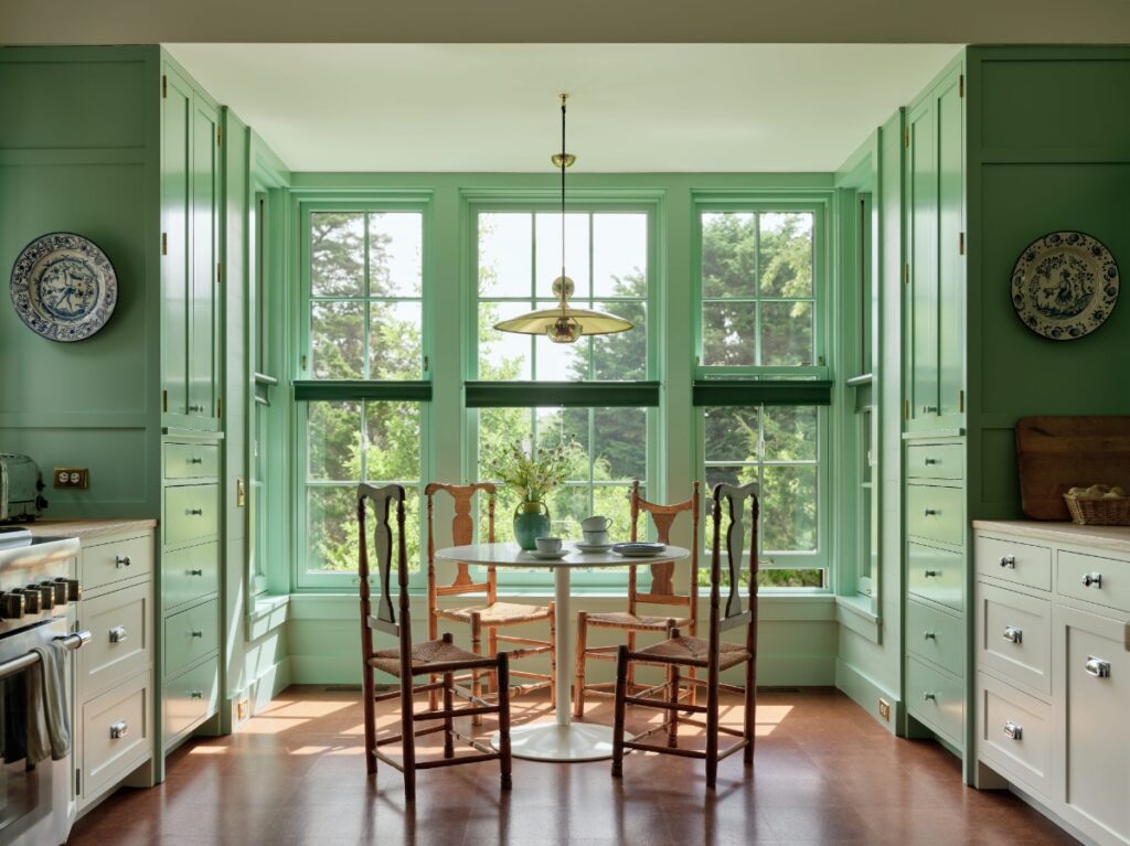 Sage green kitchen alcove with white cabinets, round dining table, woven chairs, and brass pendant light.