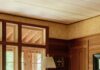 Inside a Modern Rhode Island Cottage Inspired by Childhood Memories Wood-paneled living room with wicker armchair, floor lamp, large windows, and diamond wood frieze detail.