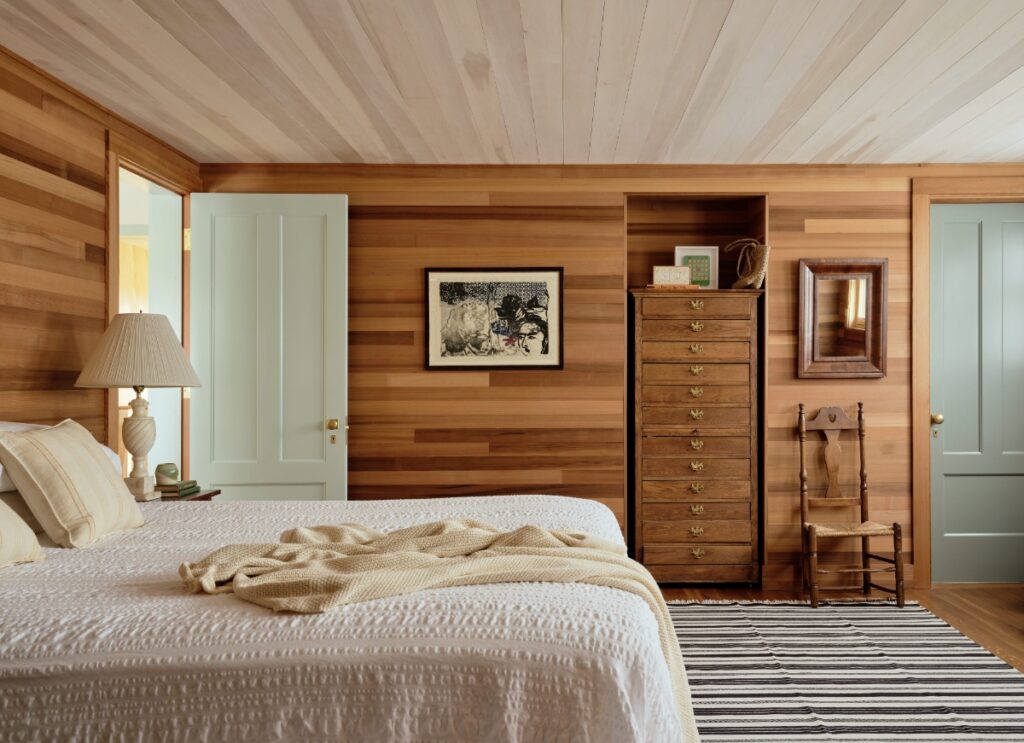 Wood-paneled bedroom featuring a white bedspread, tall wooden dresser, striped rug, and pale blue doors.