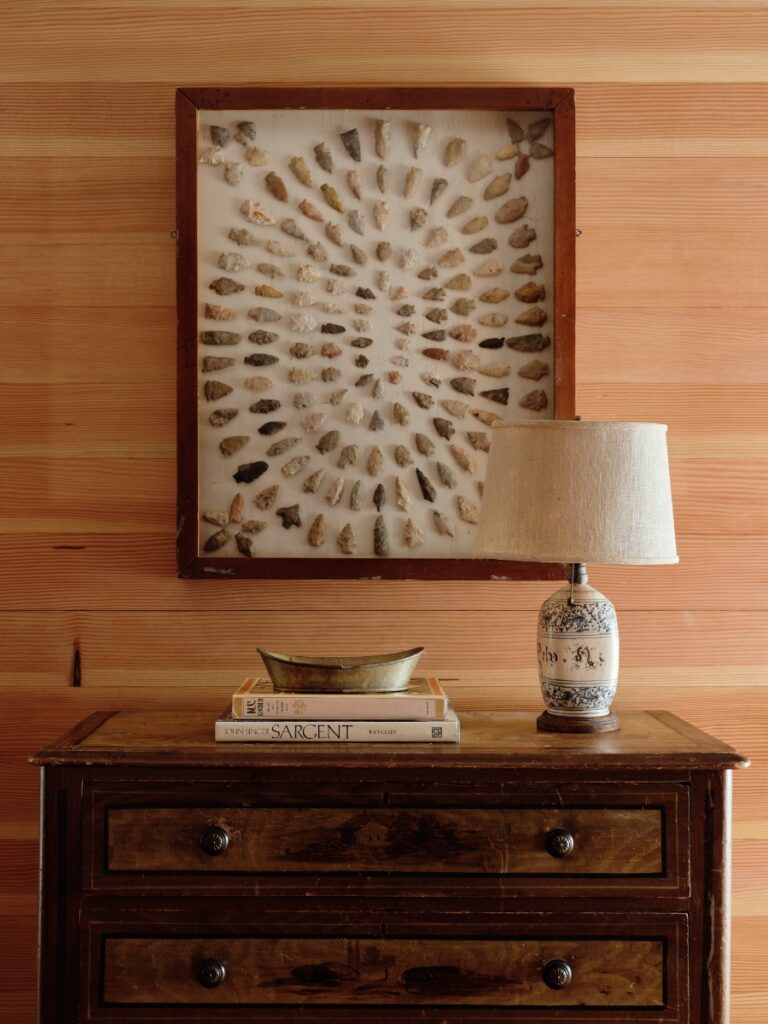 Framed arrowhead shadowbox display mounted on horizontal wood paneling above a antique wooden bureau.