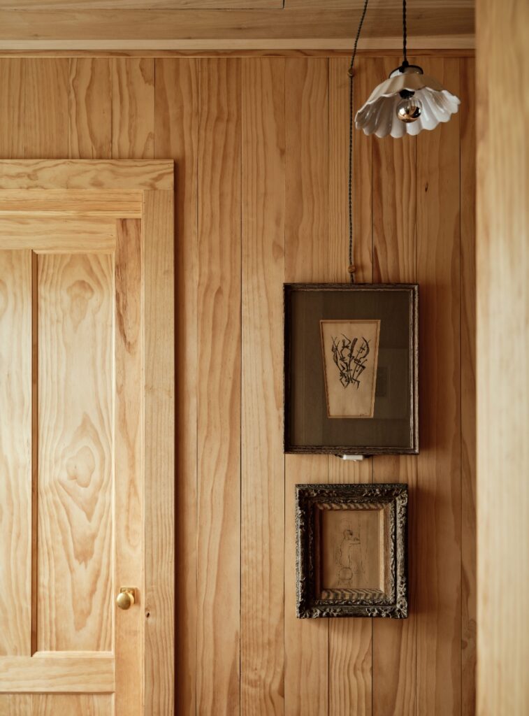 Natural pine wood paneled wall with two small framed artworks beneath a ceramic pendant lamp.