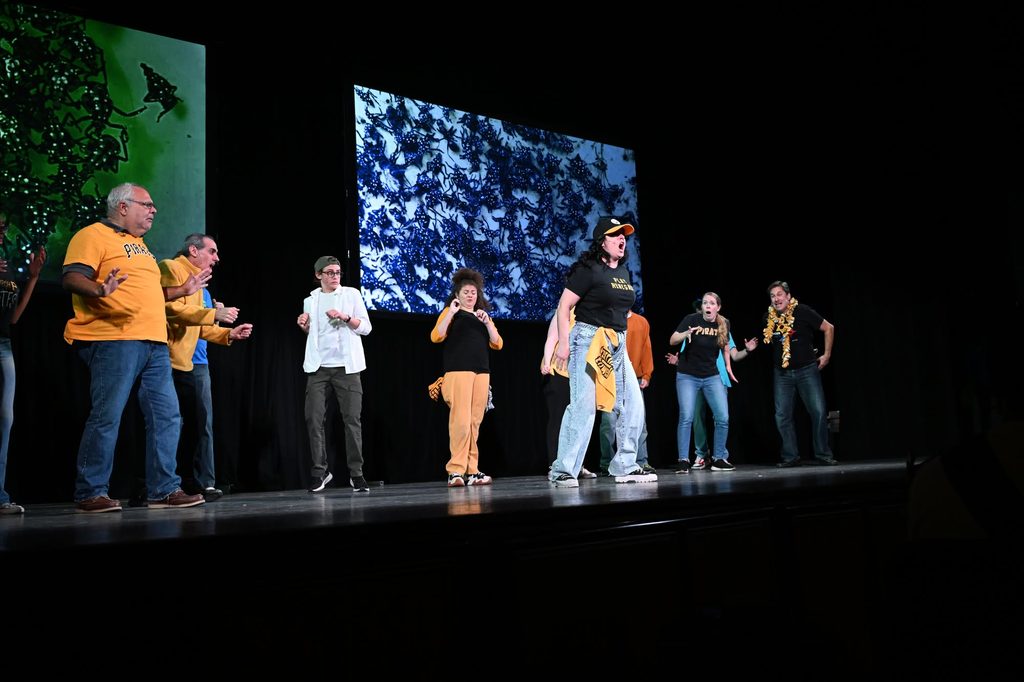 Group performing on stage in Pittsburgh, with a large screen behind them.