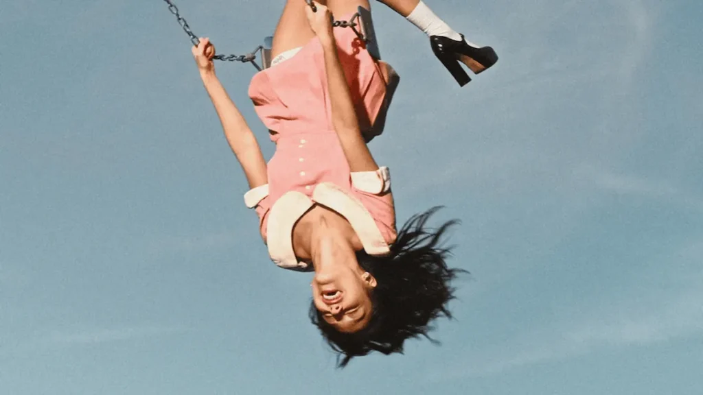 Olivia Rodrigo upside down on a swing, wearing a pink dress and platform heels.