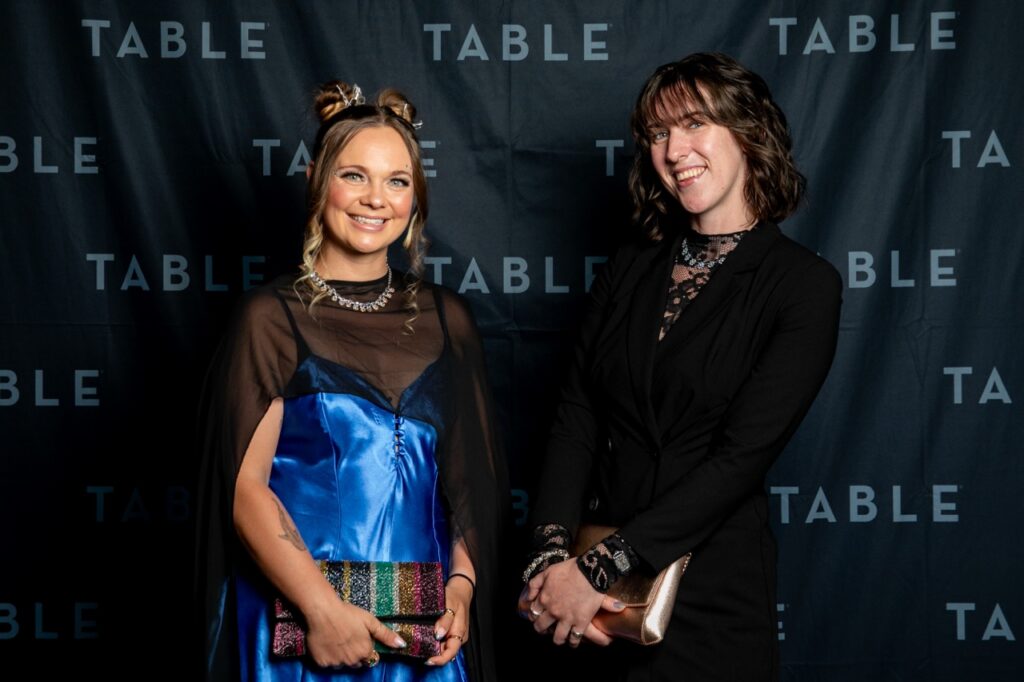 Two women in formal attire pose for a photo in front of a black backdrop with the word "TABLE" printed repeatedly.