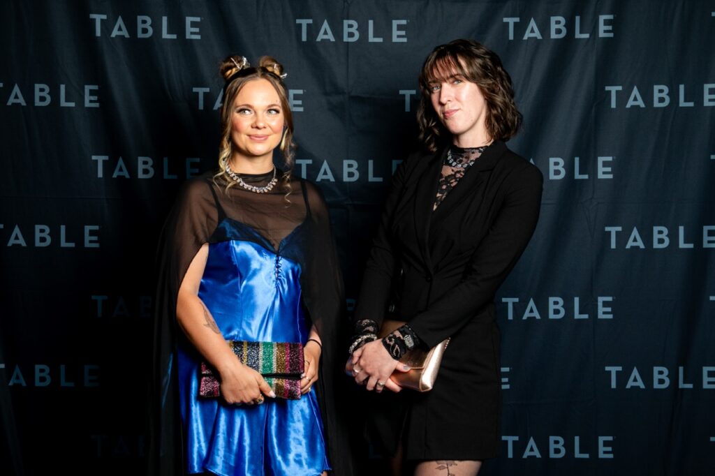 Two women in formal attire pose in front of a black backdrop with "TABLE" repeated.