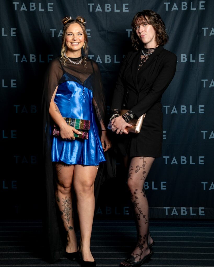 Two women in formal attire pose in front of a backdrop with the word "TABLE" repeated.