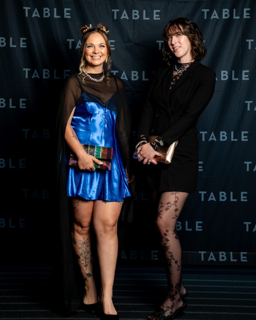 Two women in formal attire pose in front of a black backdrop with the word "TABLE" repeated