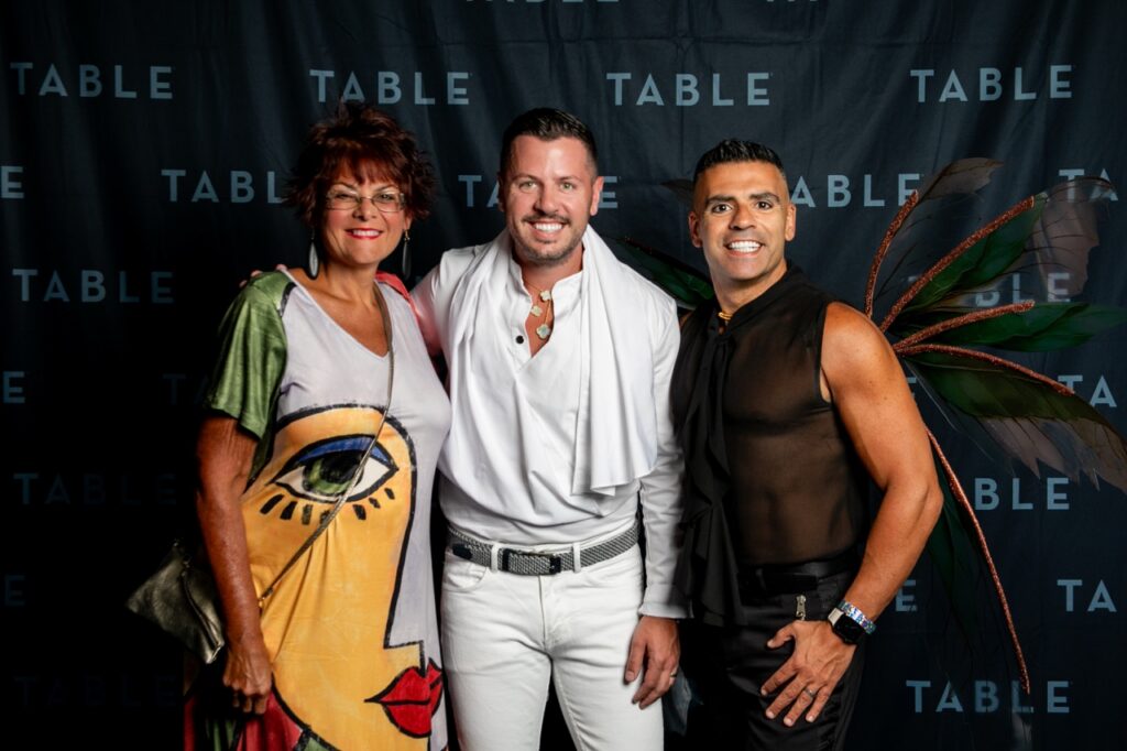 Three smiling people pose for a photo in front of a black TABLE backdrop, with one person wearing fairy wings.