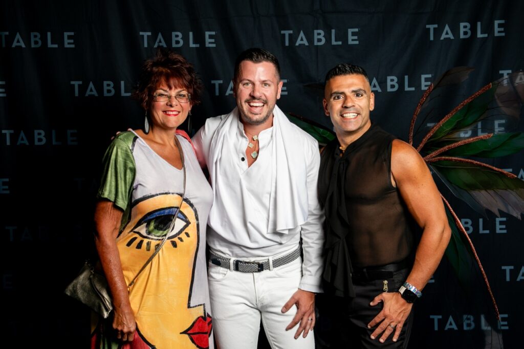 Three people pose for a photo in front of a TABLE backdrop, including a woman in a colorful dress and two men.