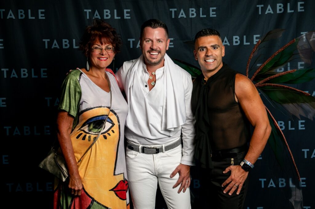 Three people pose for a photo in front of a TABLE backdrop, one with fairy wings