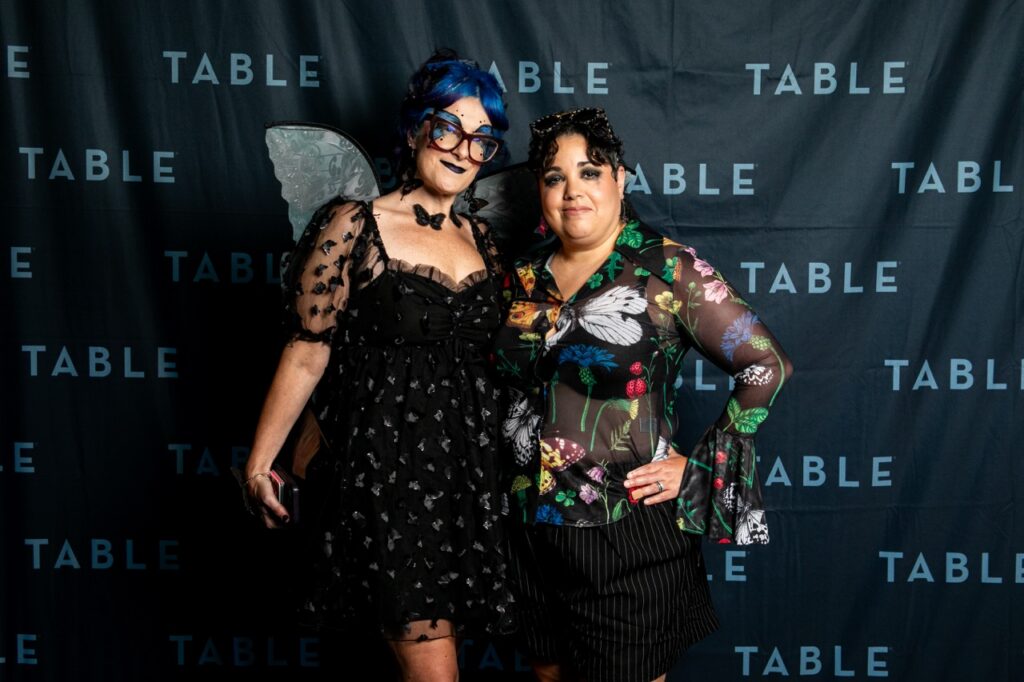 Two women in costumes pose in front of a black backdrop with "TABLE" repeatedly printed on it.
