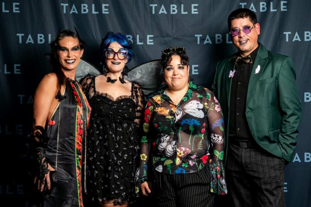 Four people in costumes pose in front of a backdrop with "TABLE" logo