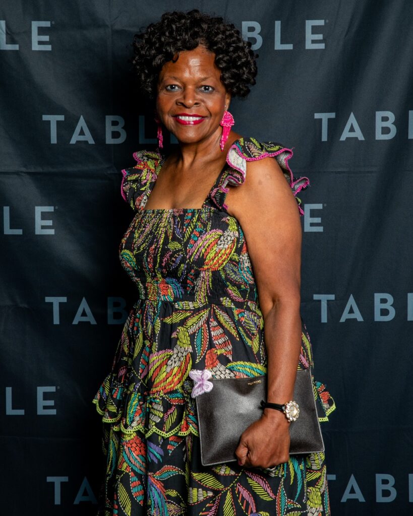 Woman in a colorful dress smiles at the camera in front of a black backdrop with "TABLE" repeated