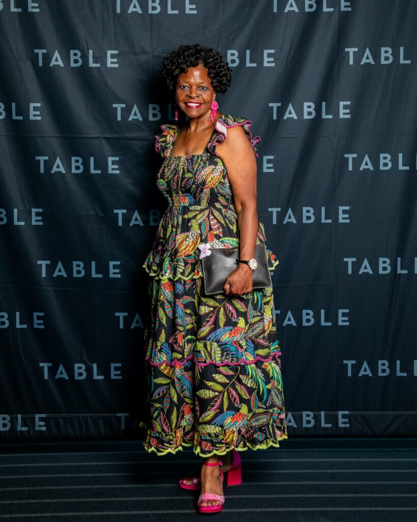Woman in a colorful floral dress and pink heels stands in front of a black backdrop with "TABLE" repeated.