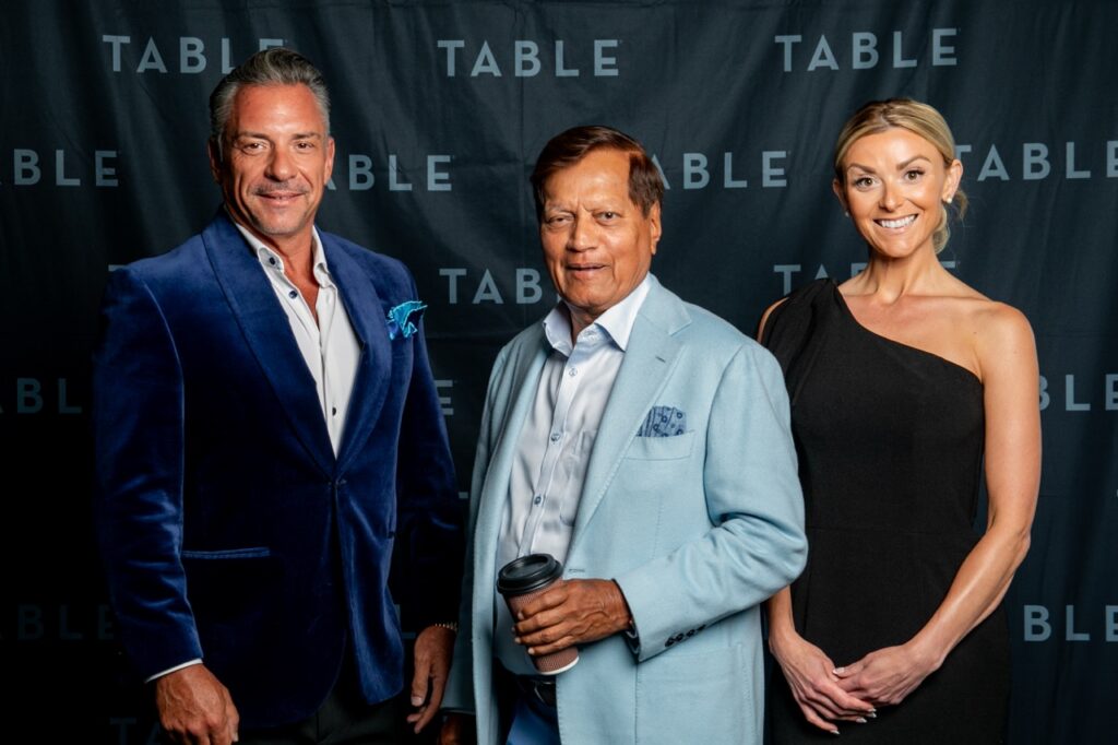 Three people pose for a photo in front of a backdrop with the word "TABLE" repeated.