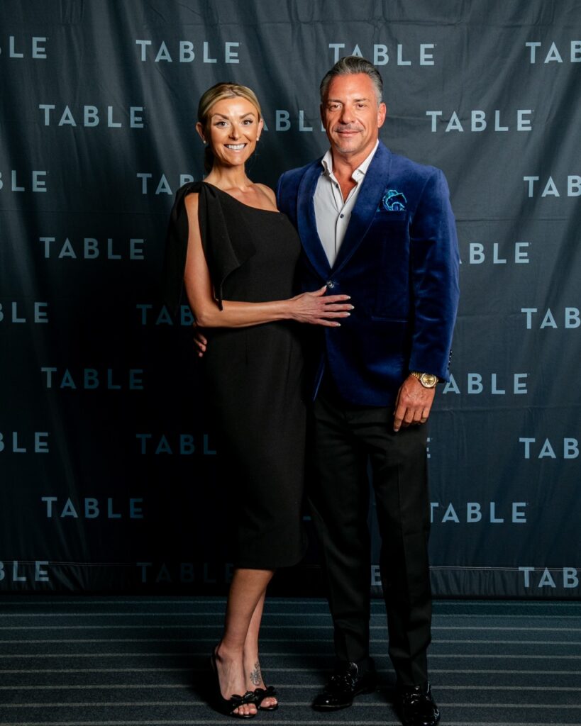 Couple poses for a photo in front of a backdrop with "TABLE" repeated