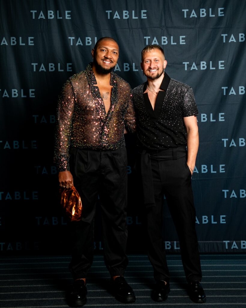 Two men in sparkling shirts and black pants pose in front of a "TABLE" backdrop.