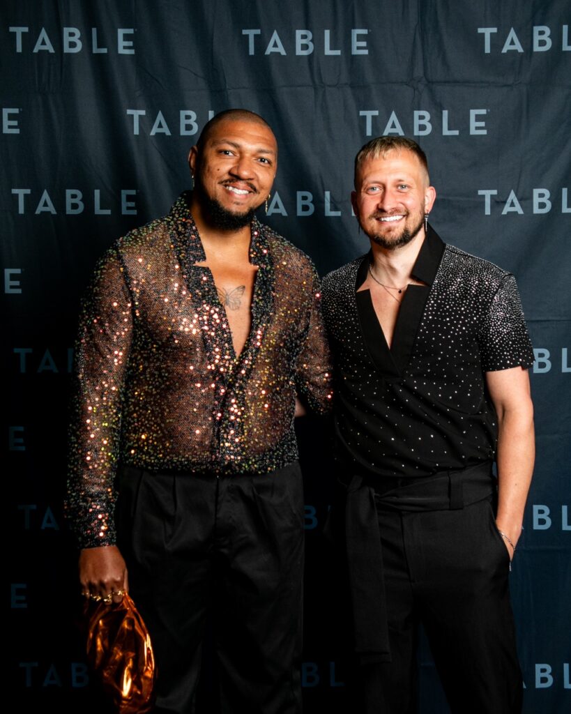 Two men in sparkly shirts pose for a photo in front of a black backdrop with the word "TABLE" repeated.