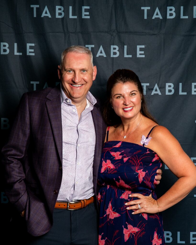 Man and woman smiling in front of a black backdrop with the word "TABLE" repeated