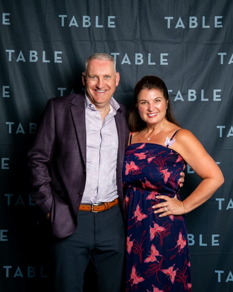Man and woman smiling in front of a TABLE backdrop at an event