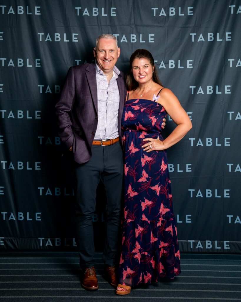 Man and woman posing in front of a TABLE backdrop at an event