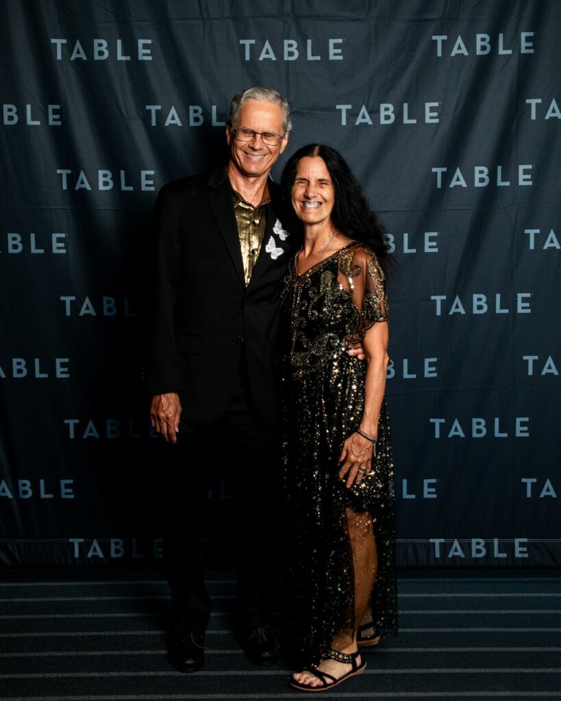 Couple in formal attire poses in front of a "TABLE" backdrop at an event.