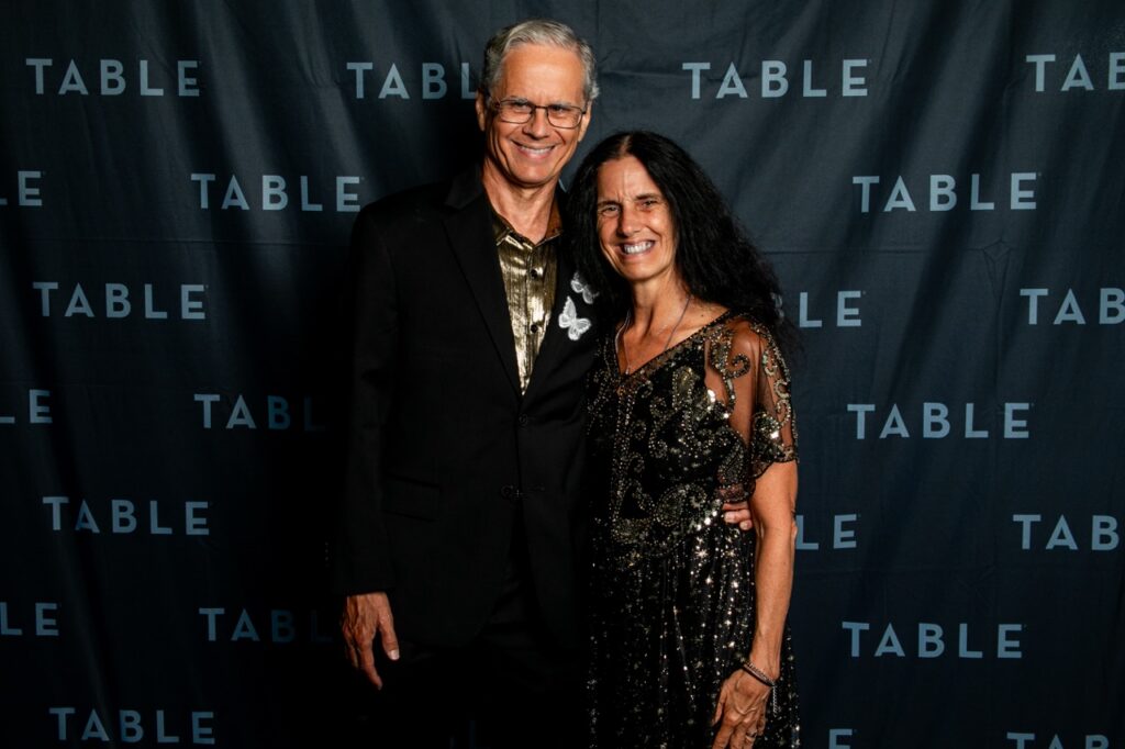 Man in black suit and woman in sparkly dress smile at event with "TABLE" backdrop
