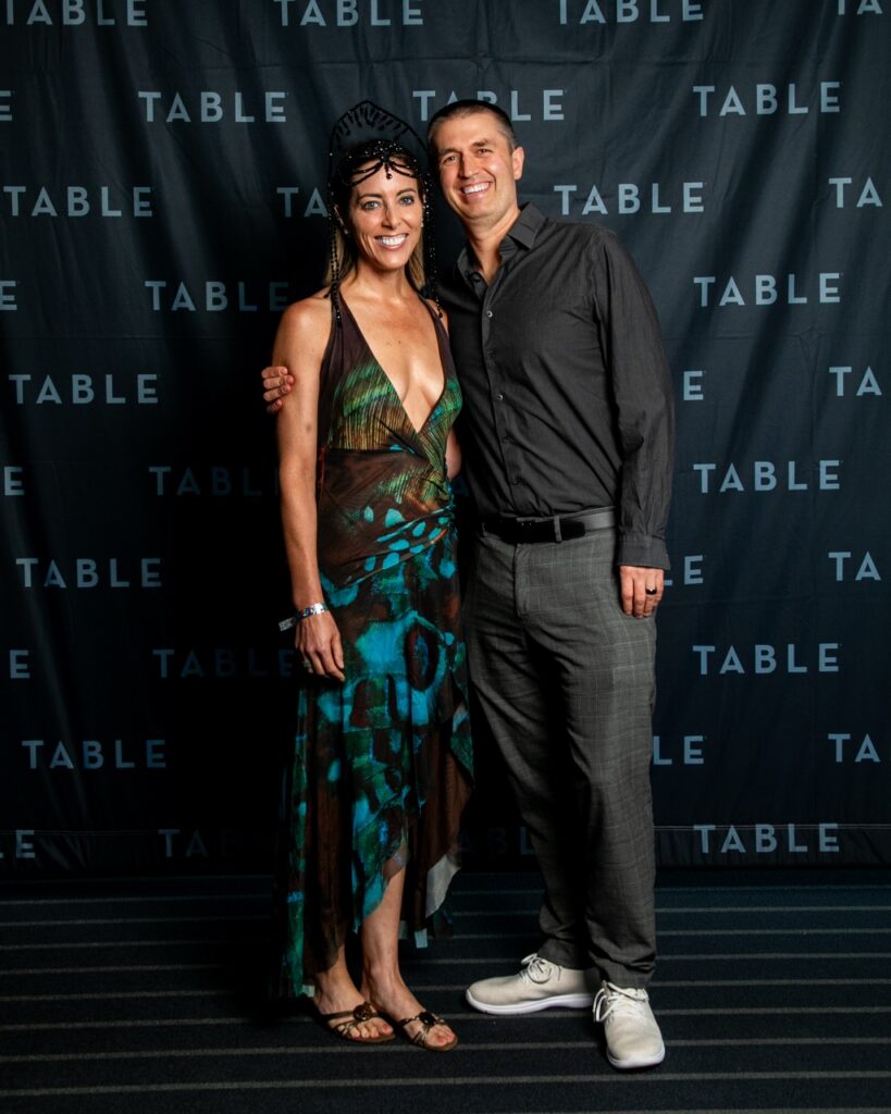 Couple poses for a photo in front of a TABLE backdrop at an event.