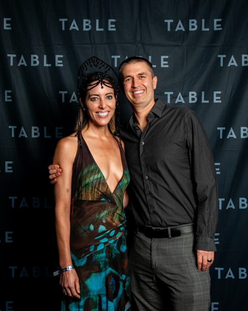 Couple posing in front of a black backdrop with the word "TABLE" repeated