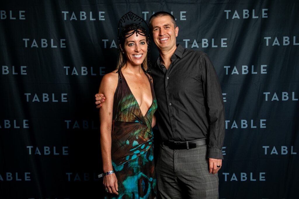 A smiling couple poses in front of a black backdrop with the word "TABLE" repeated.