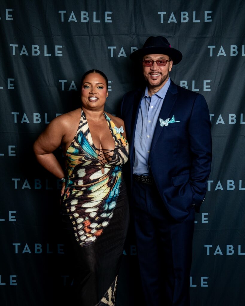 A fashionable couple poses in front of a backdrop with "TABLE" repeated.