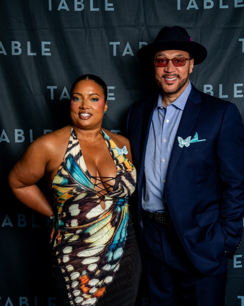 Couple in stylish outfits with butterfly accents pose in front of a black backdrop with "TABLE" logo.
