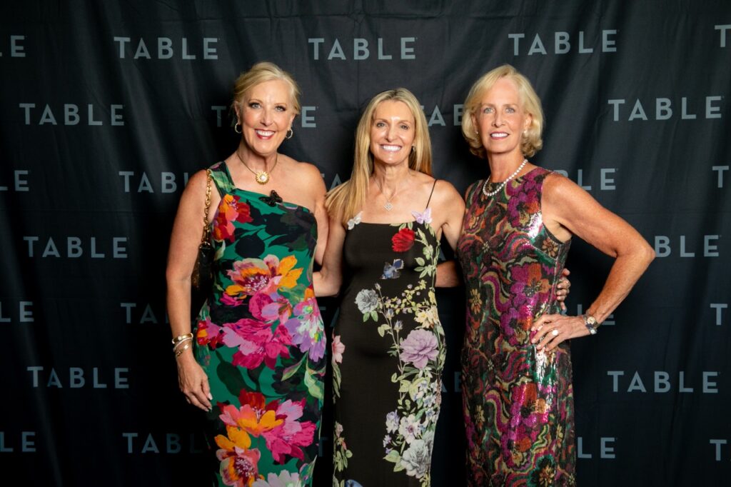Three women in colorful floral dresses pose in front of a black backdrop with "TABLE" repeated.