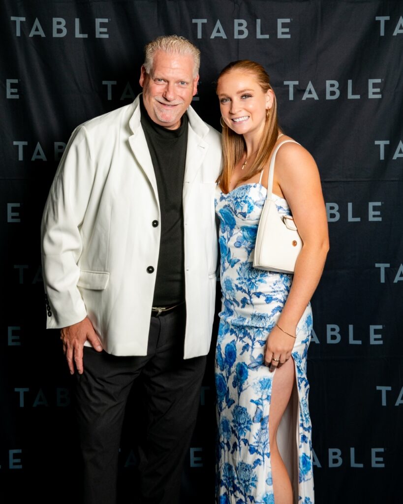 Man in white blazer and woman in blue floral dress pose at TABLE event