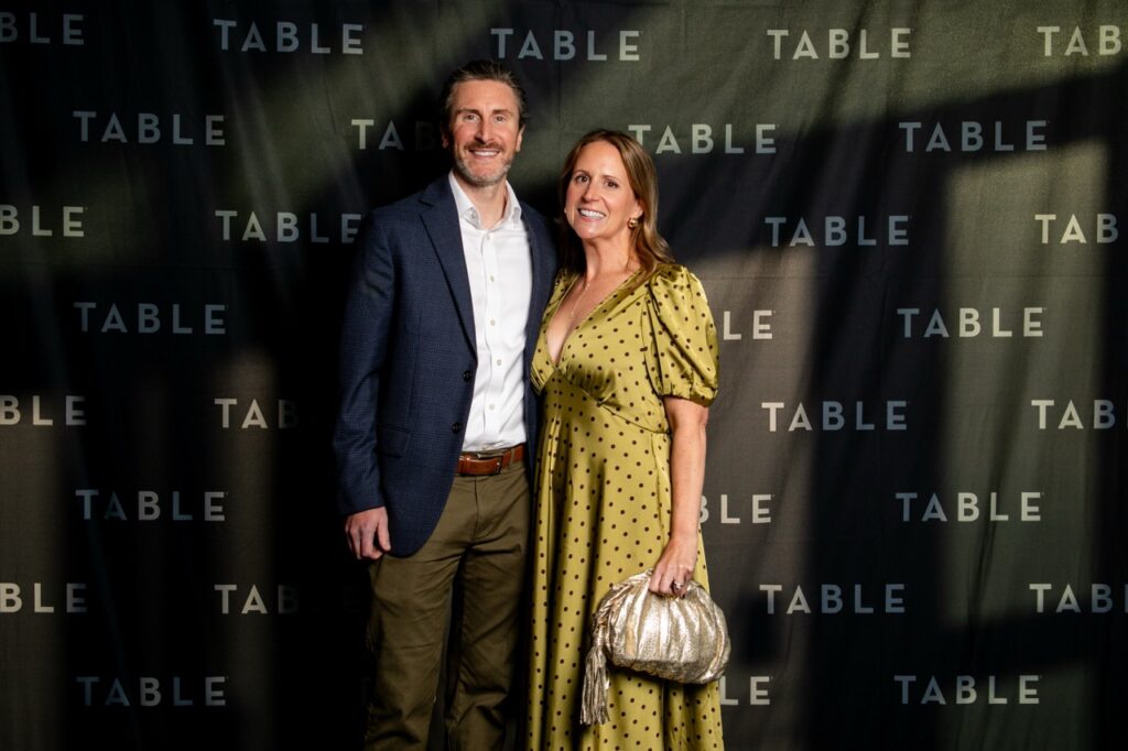 Couple poses at an event with "TABLE" repeated on the backdrop.
