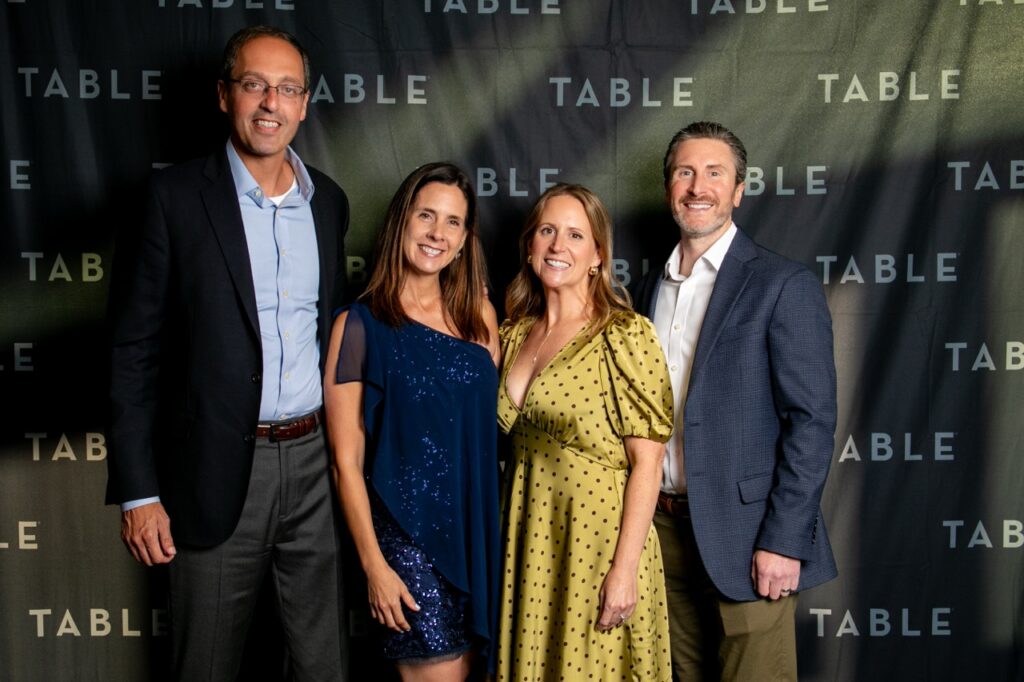 Four adults pose for a photo in front of a backdrop with the word "TABLE" repeated.