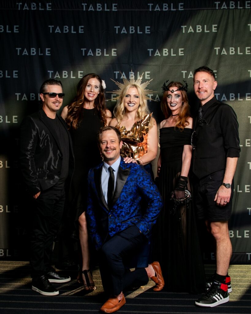 Six people in formal wear pose for a photo in front of a "TABLE" branded backdrop.