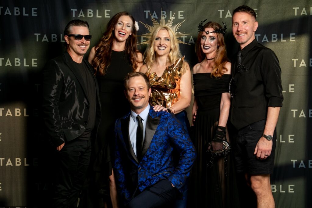 Group of six people in formal attire posing for a photo at an event with "TABLE" branding.