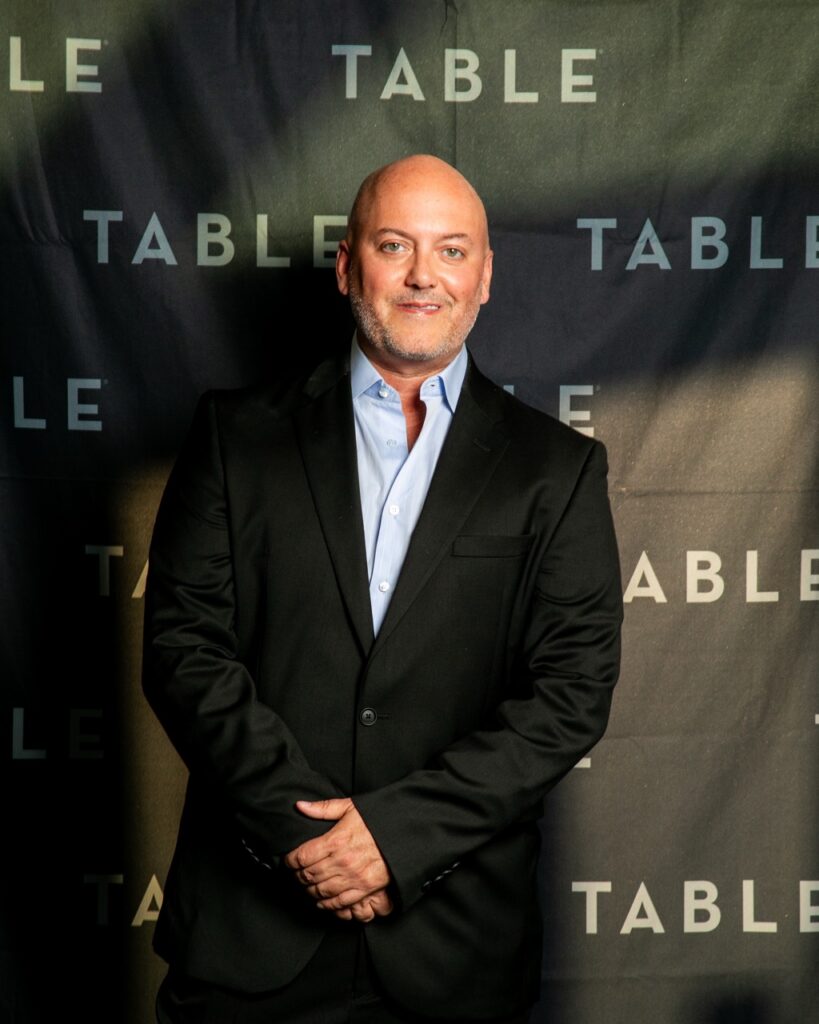 Bald man in a suit smiling in front of a TABLE backdrop