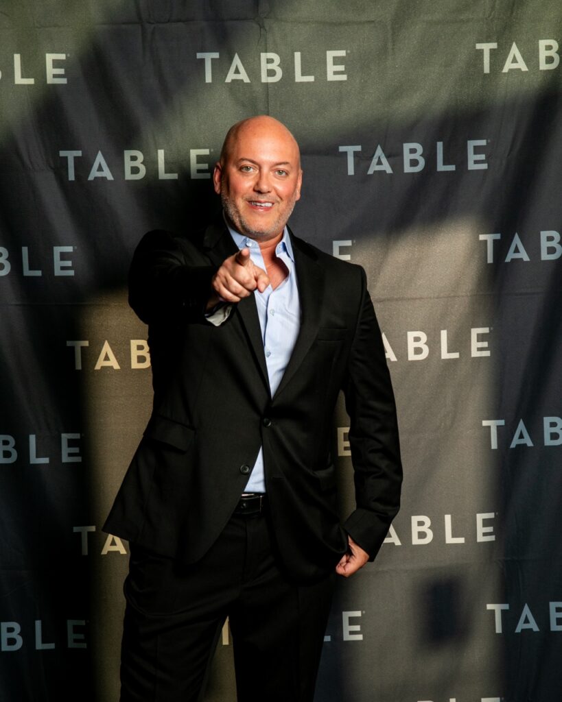 Man in a suit points at the camera in front of a "TABLE" backdrop.