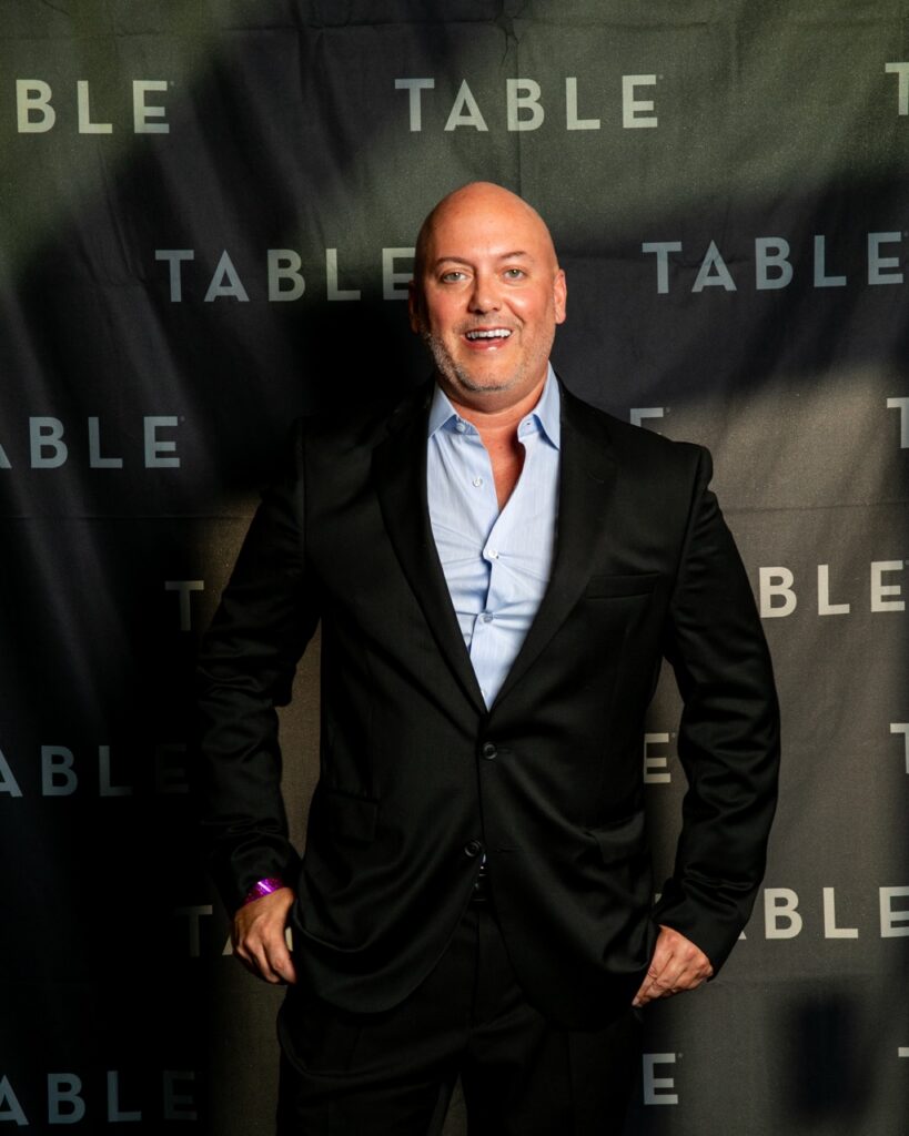 Bald man in a black suit and blue shirt smiles in front of a "TABLE" backdrop.